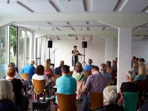 Karla Letterman, lesend im Hintergrund, im Vordergrund sitzendes Publikum